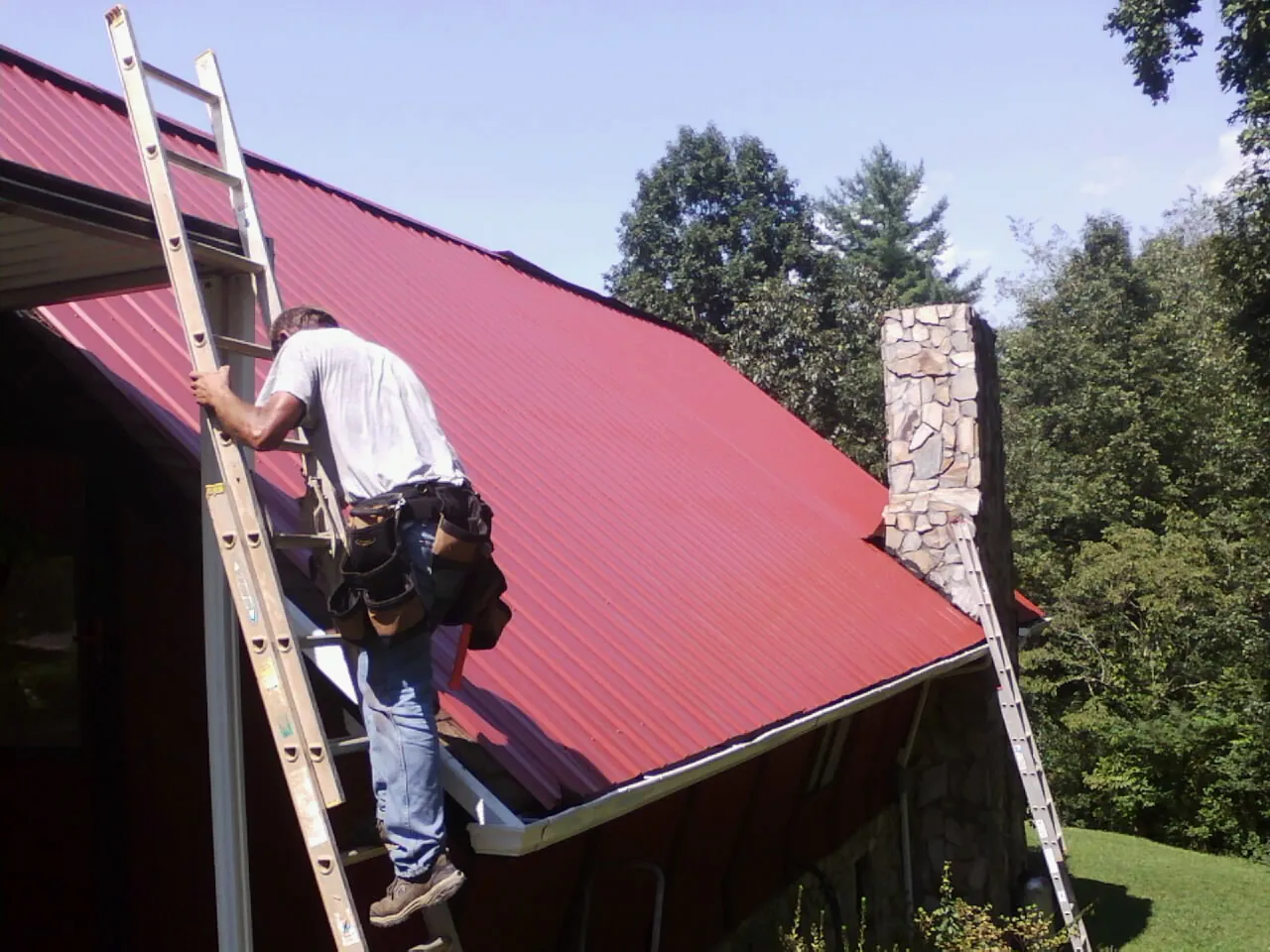 Roofer on a ladder beside a red metal roof and stone chimney — Soffit & Fascia Repair by Roofing Brothers in Hoodsport